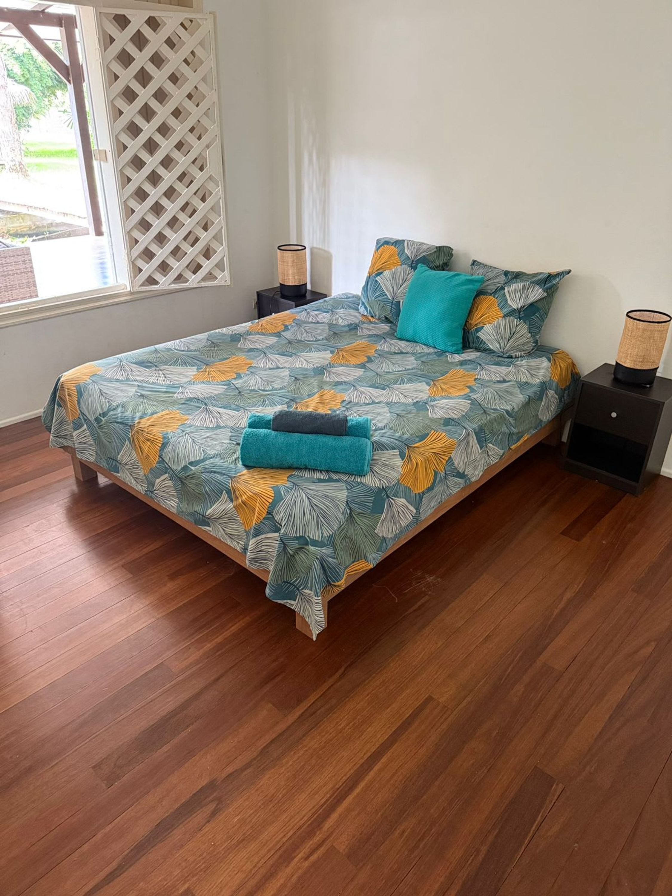 Een slaapkamer met een bed, houten vloeren, een raam met uitzicht en een nachtkastje met een lamp.