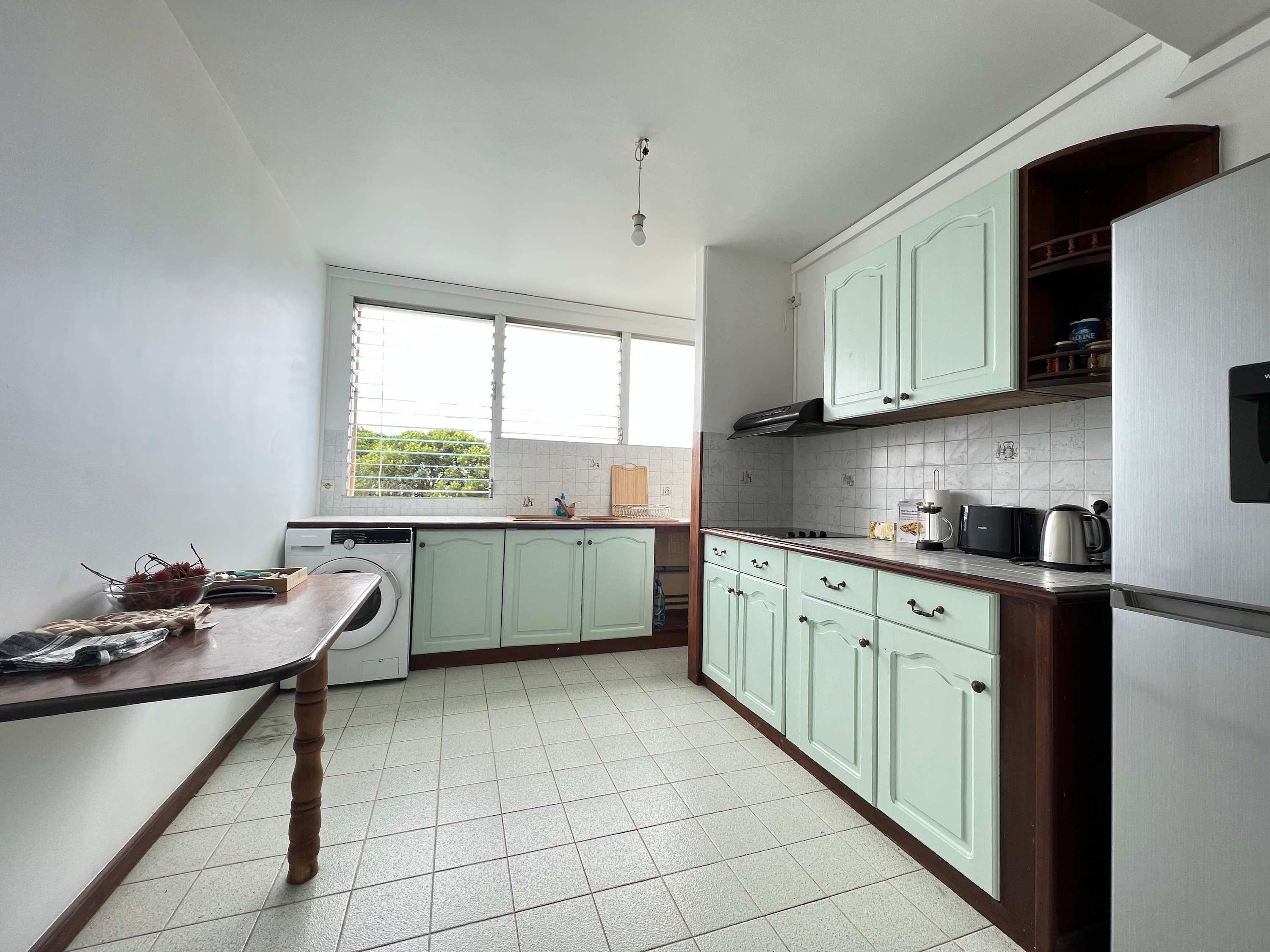 A kitchen with light green cabinets, a stainless steel refrigerator, a built-in oven, and a window with blinds.