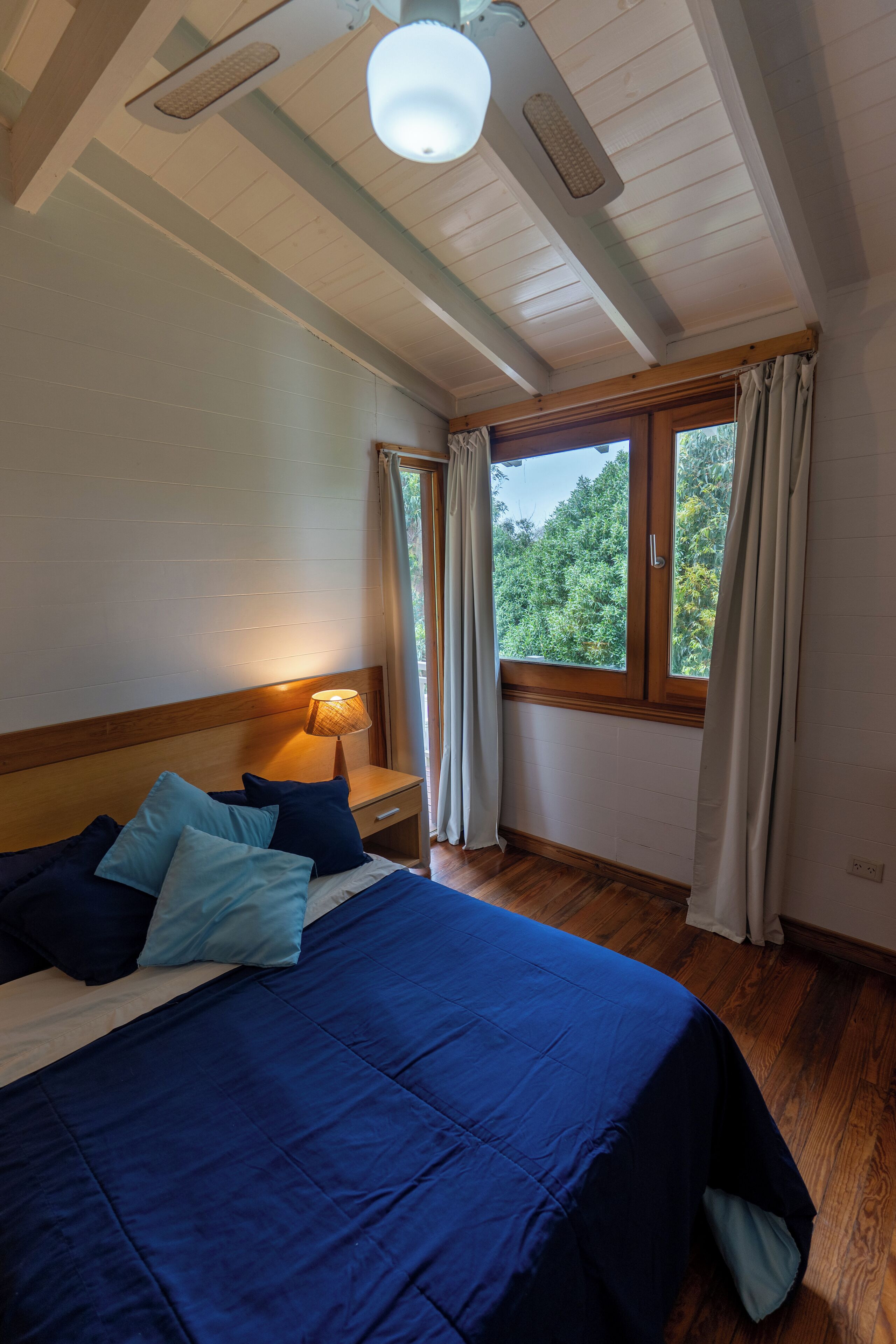 Ein Schlafzimmer mit einem großen Bett, einem Holz-Kopfteil, einem Nachttisch mit Lampe, einem Fenster mit Vorhängen und Blick ins Grüne.