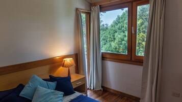 Ein Schlafzimmer mit einem großen Bett, einem Holz-Kopfteil, einem Nachttisch mit Lampe, einem Fenster mit Vorhängen und Blick ins Grüne.