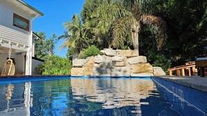 Una piscina con muro de piedra y plantas tropicales, un edificio blanco con amplios ventanales y una terraza de madera.
