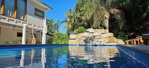 A swimming pool with a stone wall and tropical plants, a white building with large windows, and a wooden deck. - Complejo Pelicano Chapadmalal (Colonia Chapadmalal)