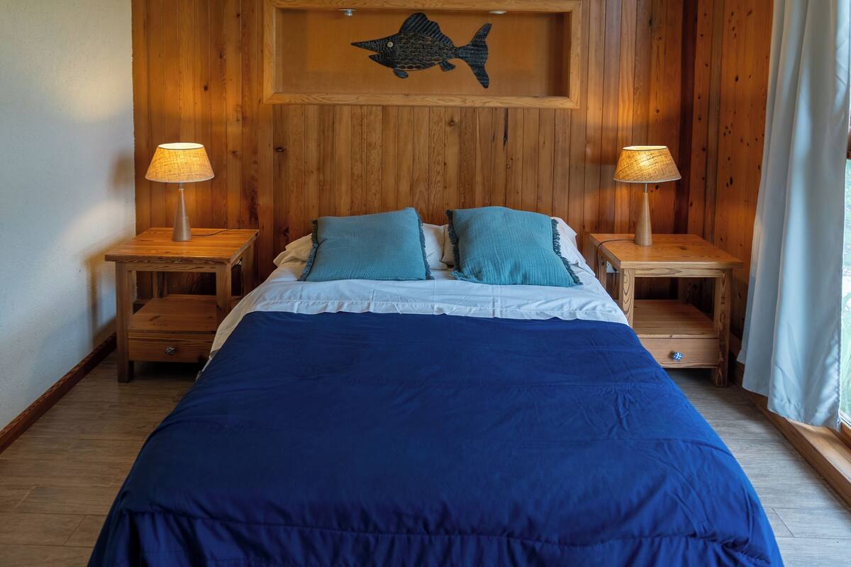 Une chambre avec un lambris en bois, un lit avec une literie bleue, deux tables de chevet avec des lampes et un luminaire au plafond.