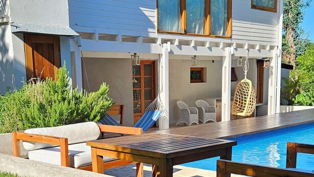 Una casa moderna con piscina, un patio con mesa y sillas, y un espacio cubierto con una silla colgante.