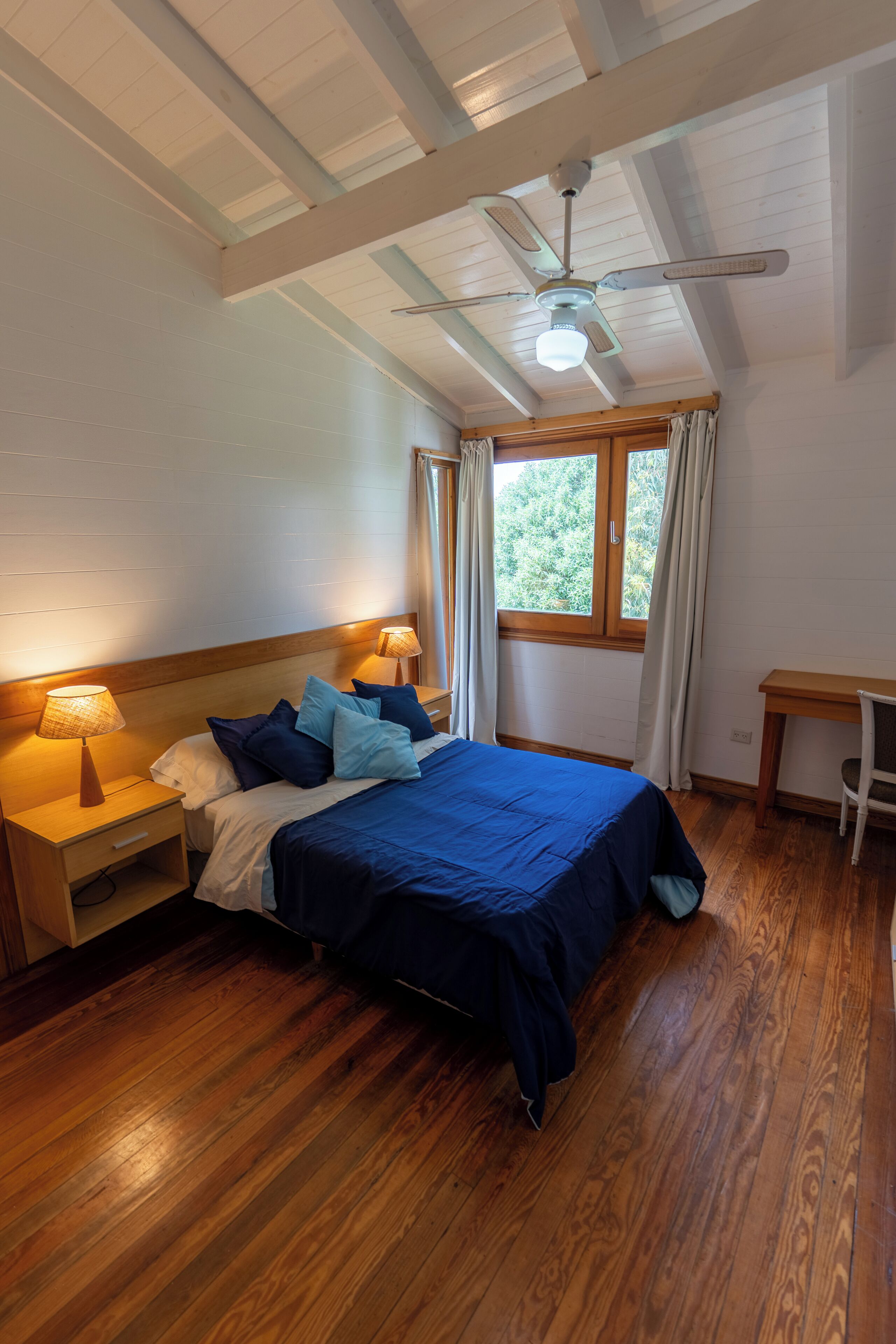 Quarto com cama, mesinhas de cabeceira de madeira, escrivaninha, ventoinha no teto e janela com cortinas.