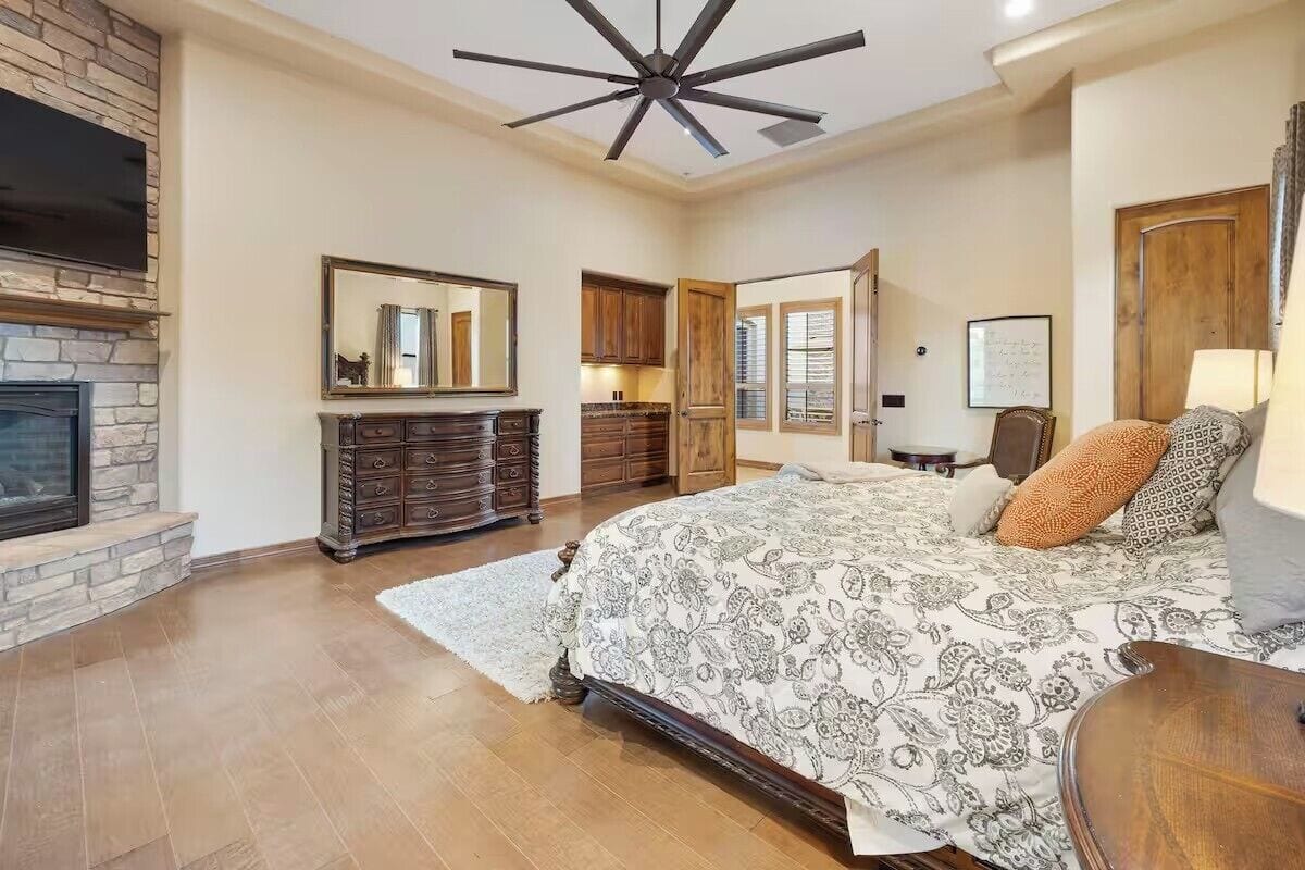 A spacious bedroom with a large bed, a fireplace, a ceiling fan, and a view of the kitchen.