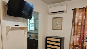 A living room with a flat-screen TV mounted on the wall, a small black table, a wicker side table, a framed picture on the wall, and a window with a view of a kitchen. - 102 Classic Double Room in waterfront hotel in Singer Island, FL (Riviera Beach)