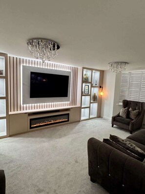 A modern living room with a large flat-screen TV, built-in shelves, a fireplace, and a chandelier. - Luxurious Majestic 4 Bedroom- Free Parking (Swanscombe)