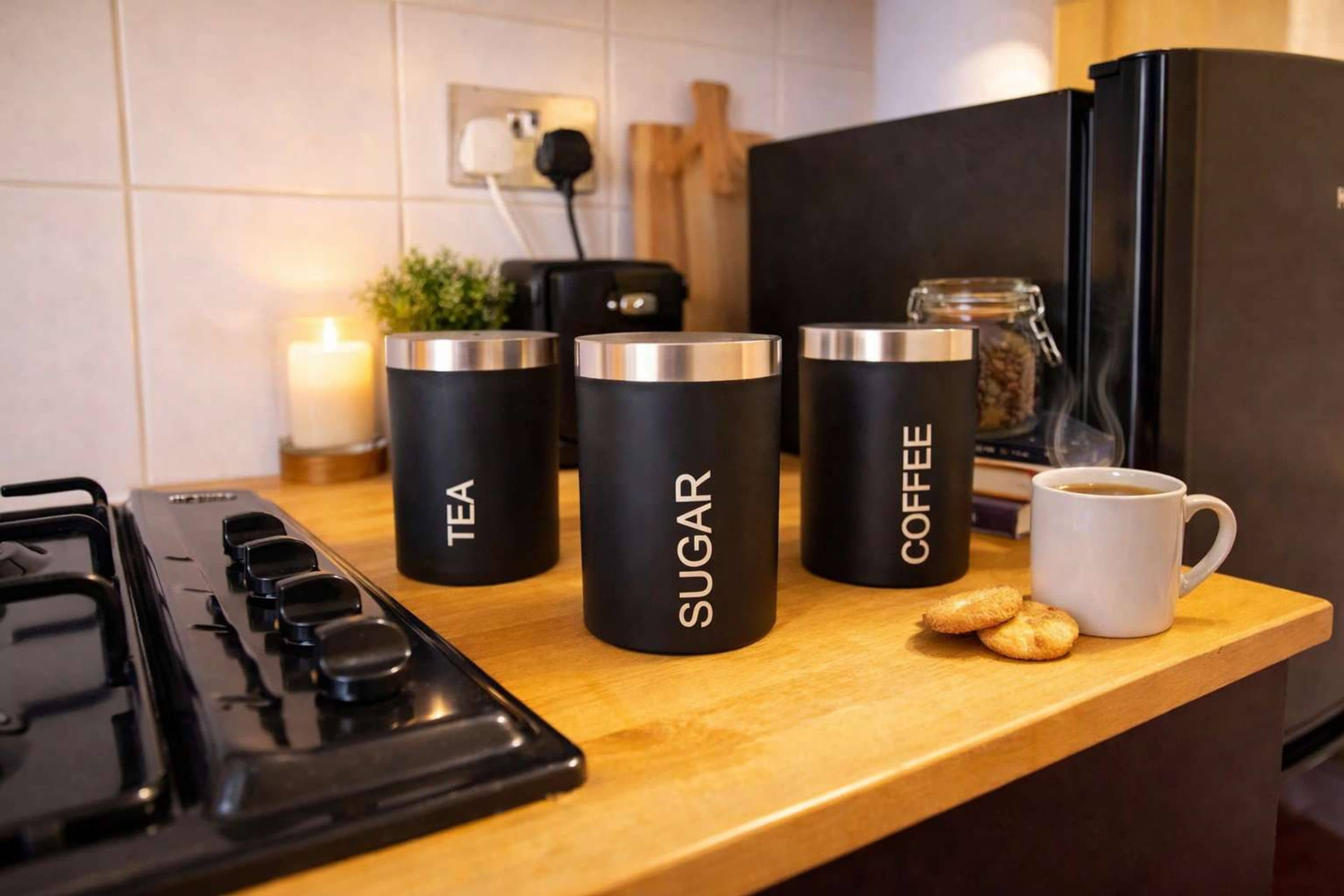 A kitchen counter with three black containers labeled 'TEA,' 'SUGAR,' and 'COFFEE,' a white mug with coffee, and a small plant.