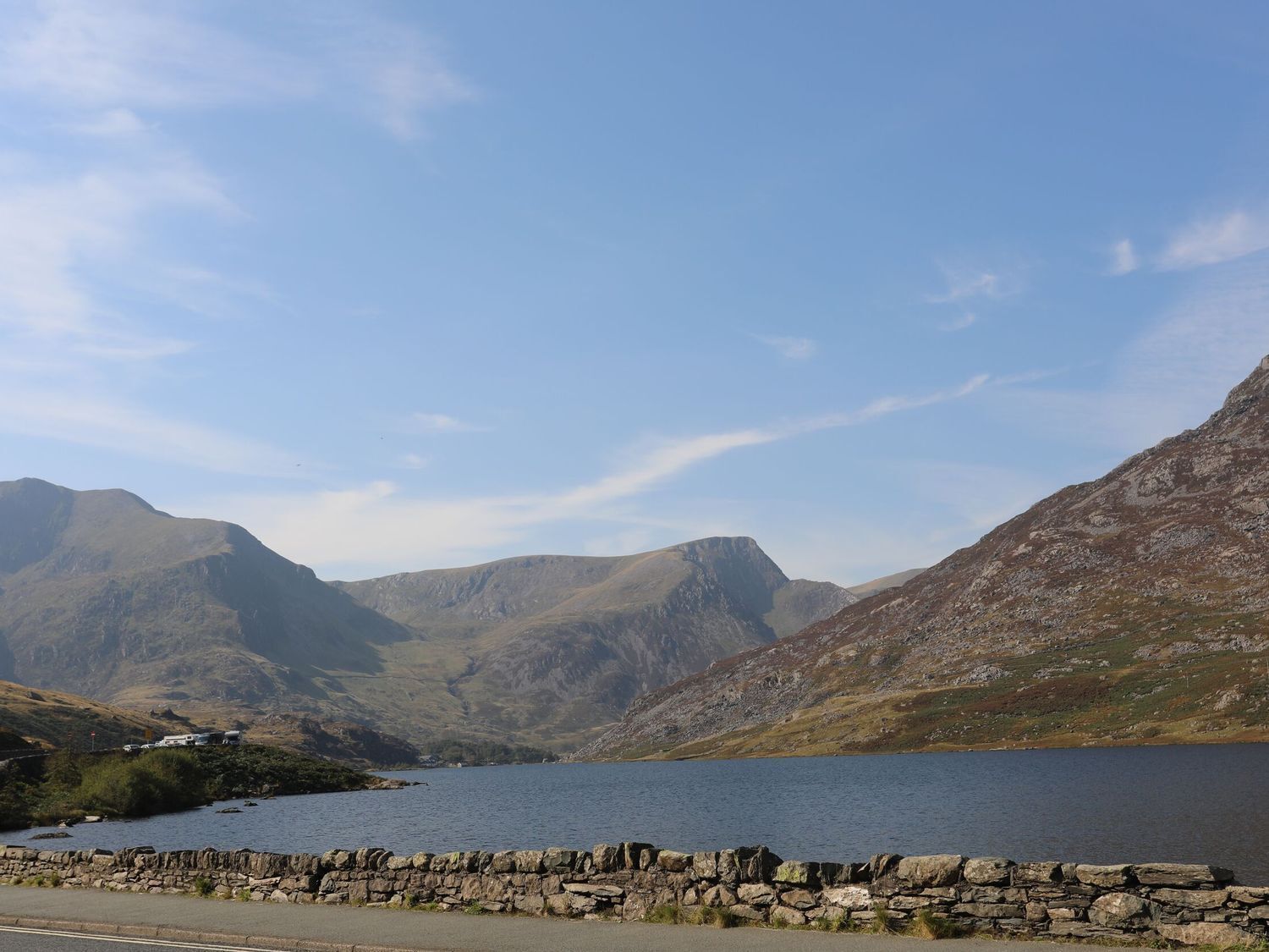 A stone wall along a road with a lake and mountains in the background.