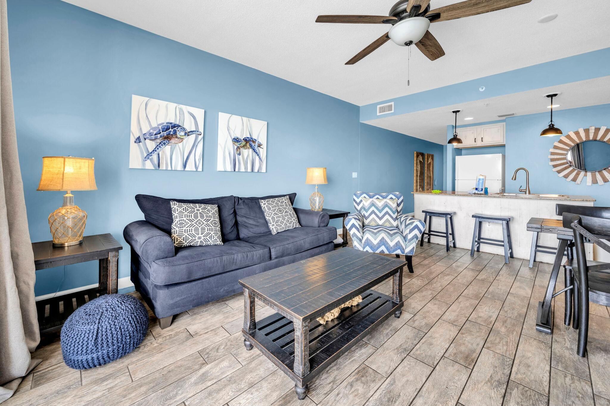 A living room with a dark blue sofa, two side tables with lamps, a coffee table, and a blue pouf. There is a ceiling fan, a kitchen area with a bar stool, and a round mirror on the wall.
