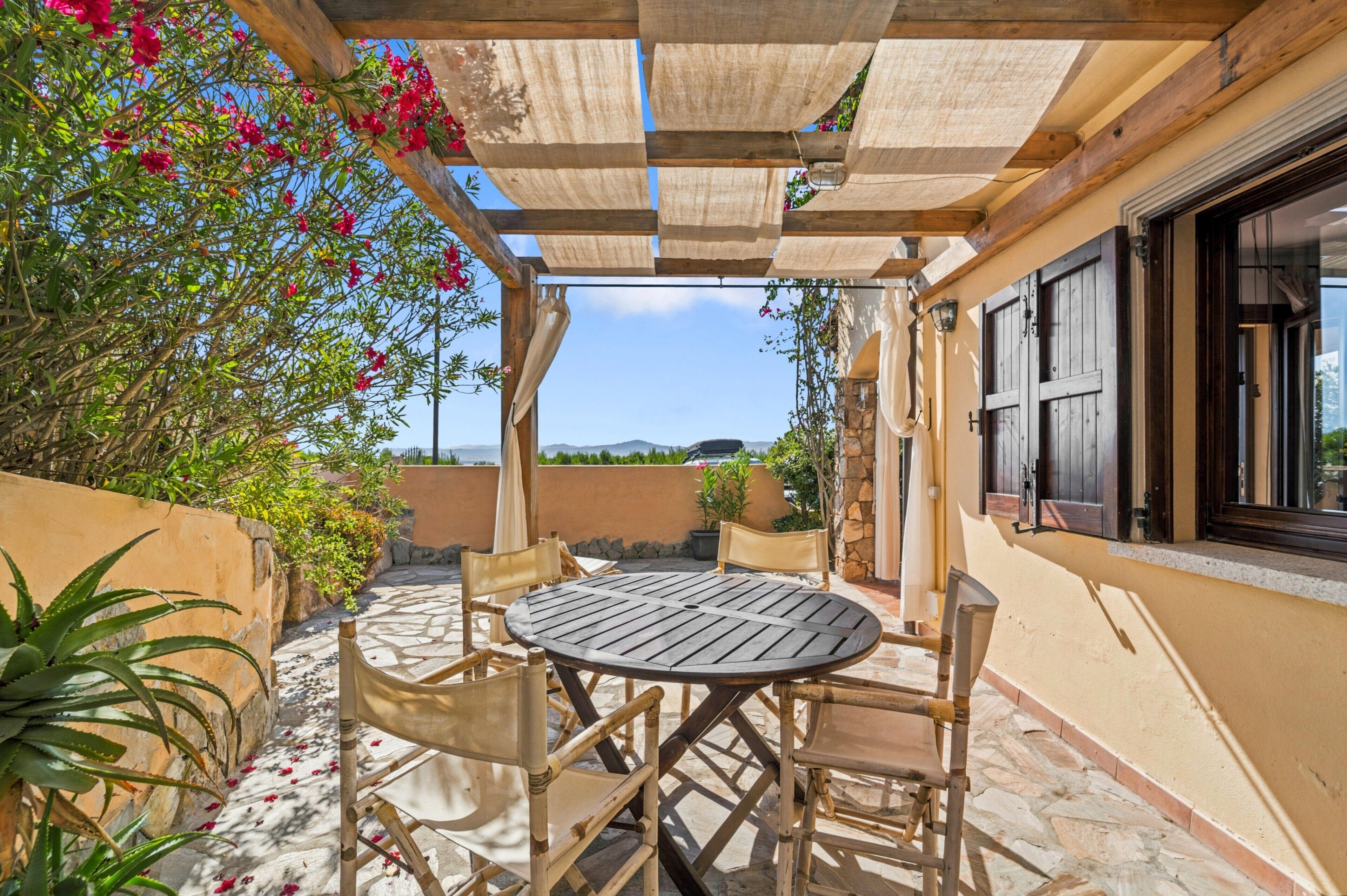 An outdoor patio with a round wooden table and chairs, surrounded by plants and a view of the landscape.