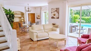 A spacious living area with a sofa, a coffee table, and a dining table with chairs. There is a staircase on the left and a sliding glass door leading to an outdoor area with a pool. - CASA-Noel villa avec piscine privée (Saint-Paul-de-Vence)
