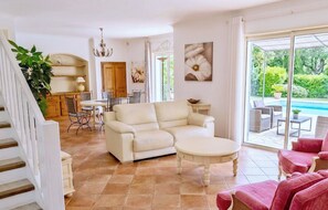 A spacious living area with a sofa, a coffee table, and a dining table with chairs. There is a staircase on the left and a sliding glass door leading to an outdoor area with a pool. - CASA-Noel villa avec piscine privée (Saint-Paul-de-Vence)