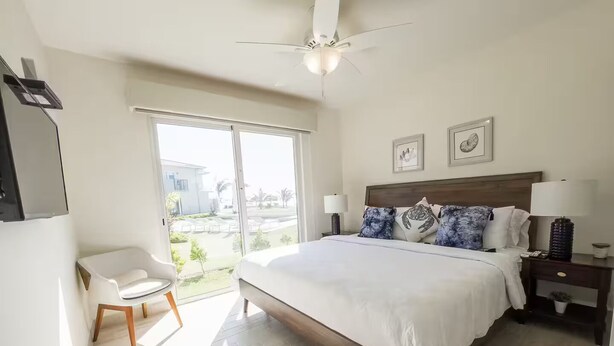Une chambre avec un grand lit, une chaise, une table de chevet, un ventilateur de plafond et une vue sur l’extérieur.