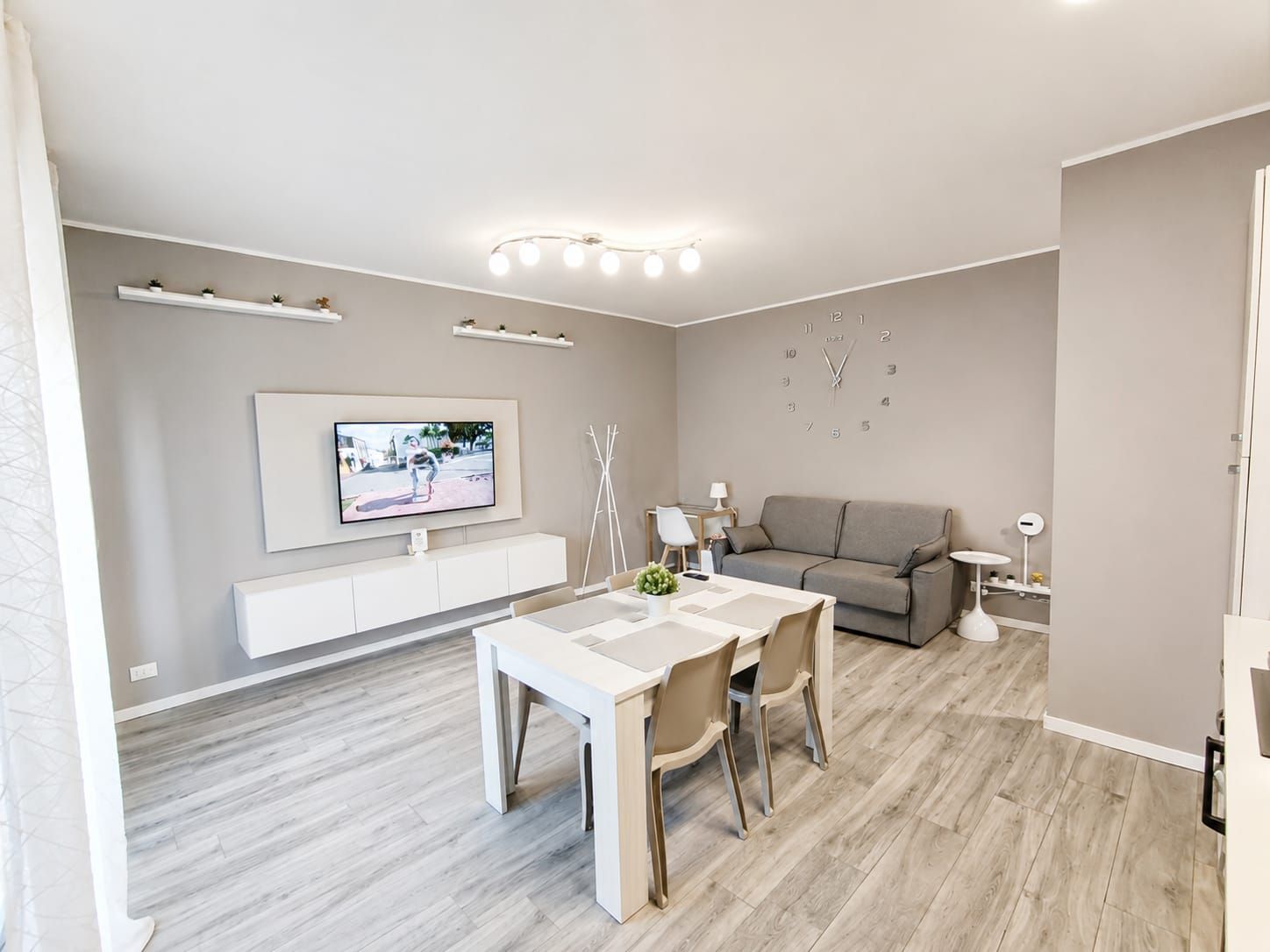 A modern living room with a dining area, a grey sofa, a flat-screen TV mounted on the wall, and a minimalist design.