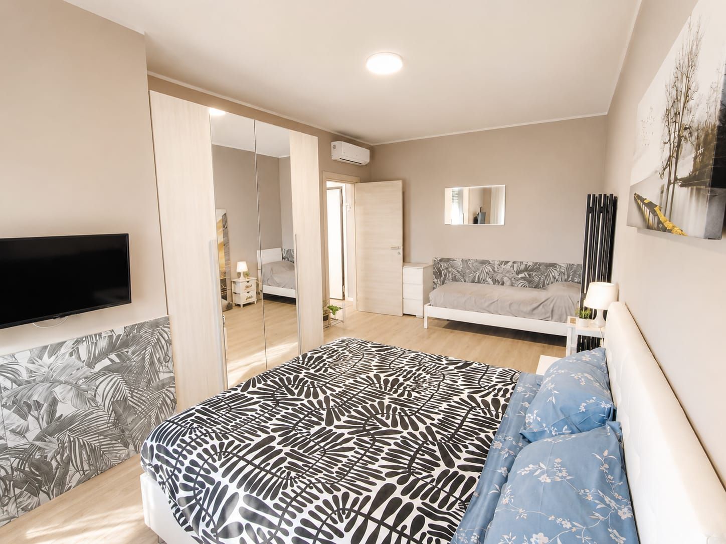 A bedroom with a bed, a TV, a sofa, a mirror, and a wall-mounted air conditioner.