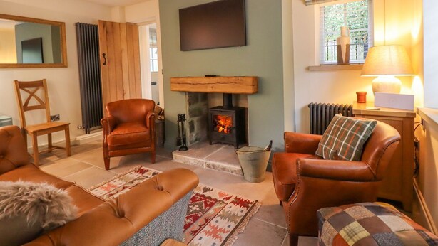 A cozy living room with a fireplace, leather sofas, a television, and a window with curtains. - Bridge House Cottage (Harrogate)