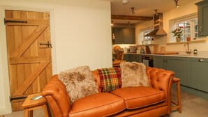 A cozy living room with a leather sofa, a wooden door, and a view into a kitchen with green cabinets and a window. - Bridge House Cottage (Harrogate)