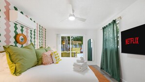 Habitación de hotel con cama, almohadas, aire acondicionado de pared, ventilador de techo, televisor mostrando 'NETFLIX' y vista al exterior a través de una puerta de vidrio.