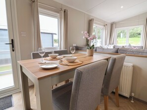 A dining area with a wooden table, grey upholstered chairs, a vase with flowers, and a view of a garden through a window. - The Oaks 2 Bedrooms (Northwich)