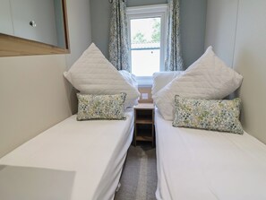A small, narrow room with two single beds, each with a pillow and a quilted headboard. A window with curtains is visible, and a small wooden shelf is placed between the beds. - The Oaks 2 Bedrooms (Northwich)