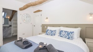 A bedroom with a large bed, a wooden beam ceiling, a round wall clock, and a bathroom visible through an open door. - Yr Hen Felin (Dyffryn Ardudwy)