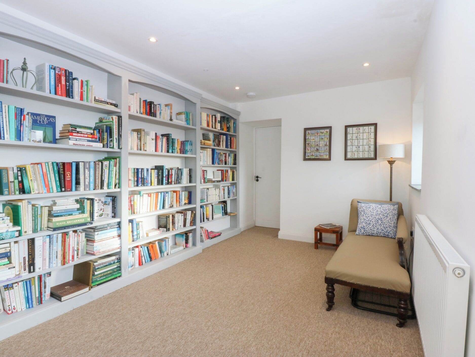 A spacious room with a large bookshelf filled with books, a small table with a lamp, and a comfortable chair.