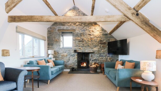 A cozy living room with a stone fireplace, two blue sofas, and a television mounted on the wall. - Yr Hen Felin (Dyffryn Ardudwy)
