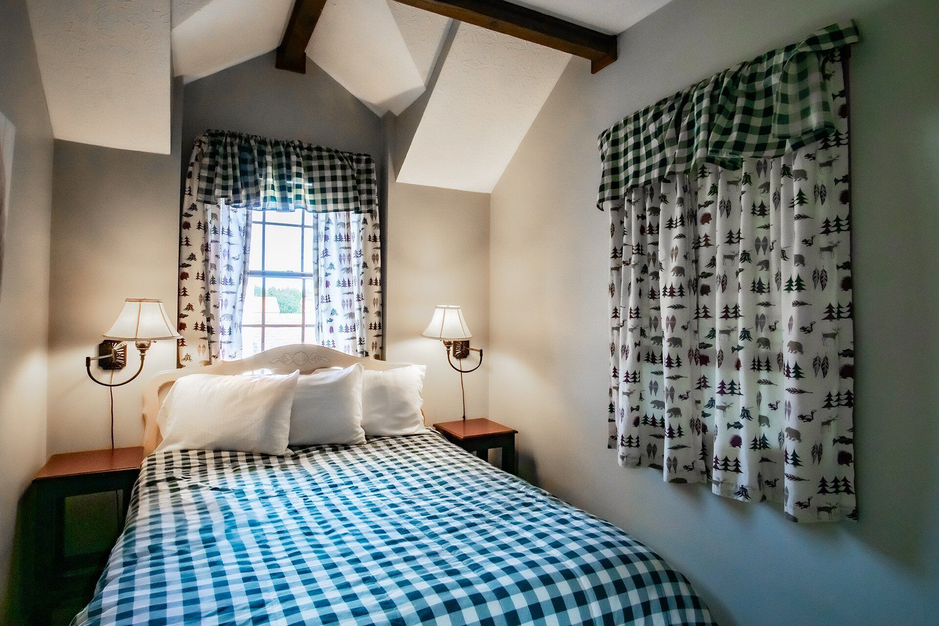 A bedroom with a checkered bedspread, two bedside lamps, a window with patterned curtains, and a wall with forest-themed curtains.