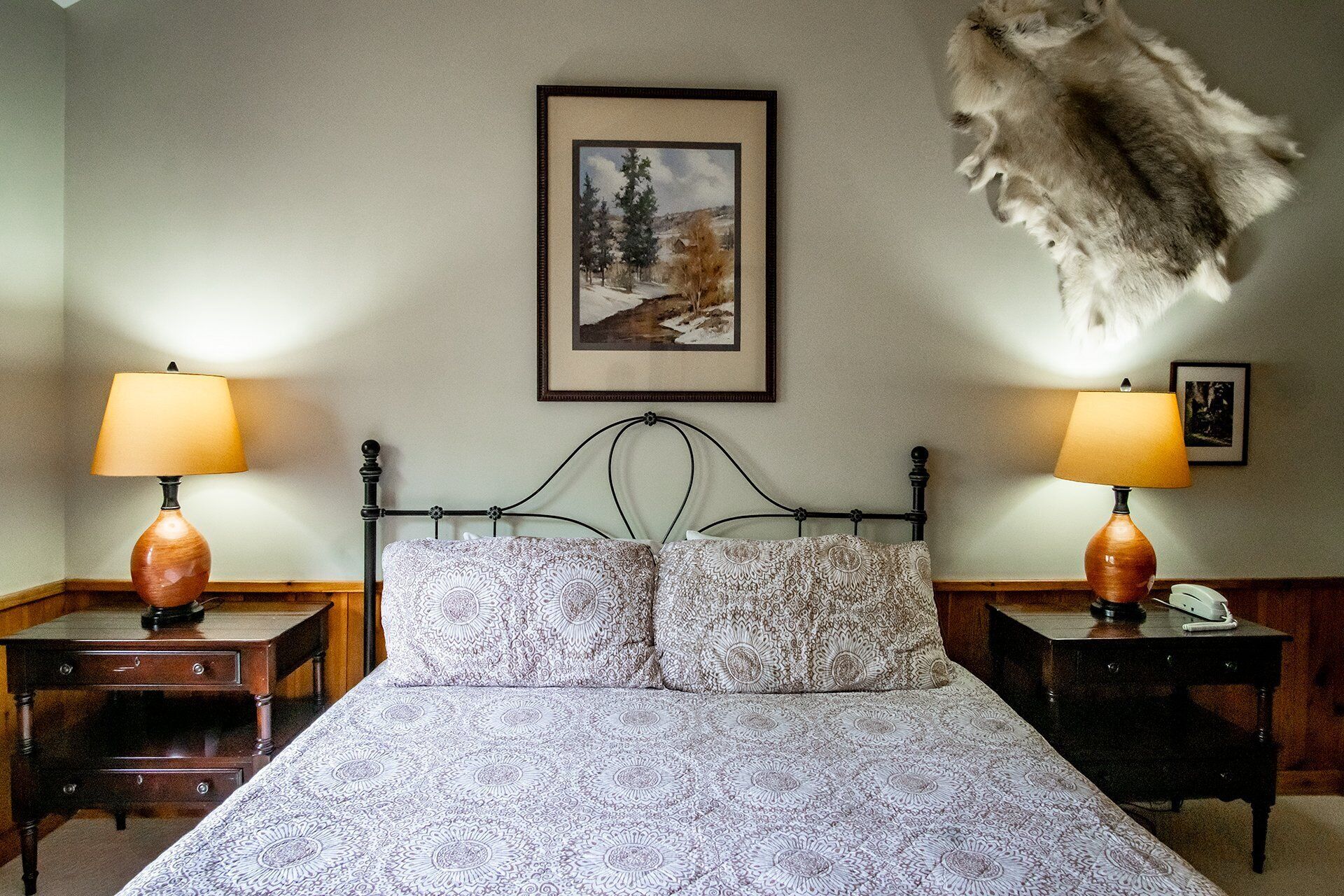 A bedroom with a bed, two bedside tables, two lamps, a framed picture on the wall, and a mounted animal head.