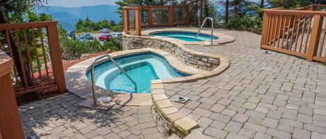 A pool area with two hot tubs, surrounded by stone walls and wooden railings, set against a backdrop of trees and a mountainous landscape.