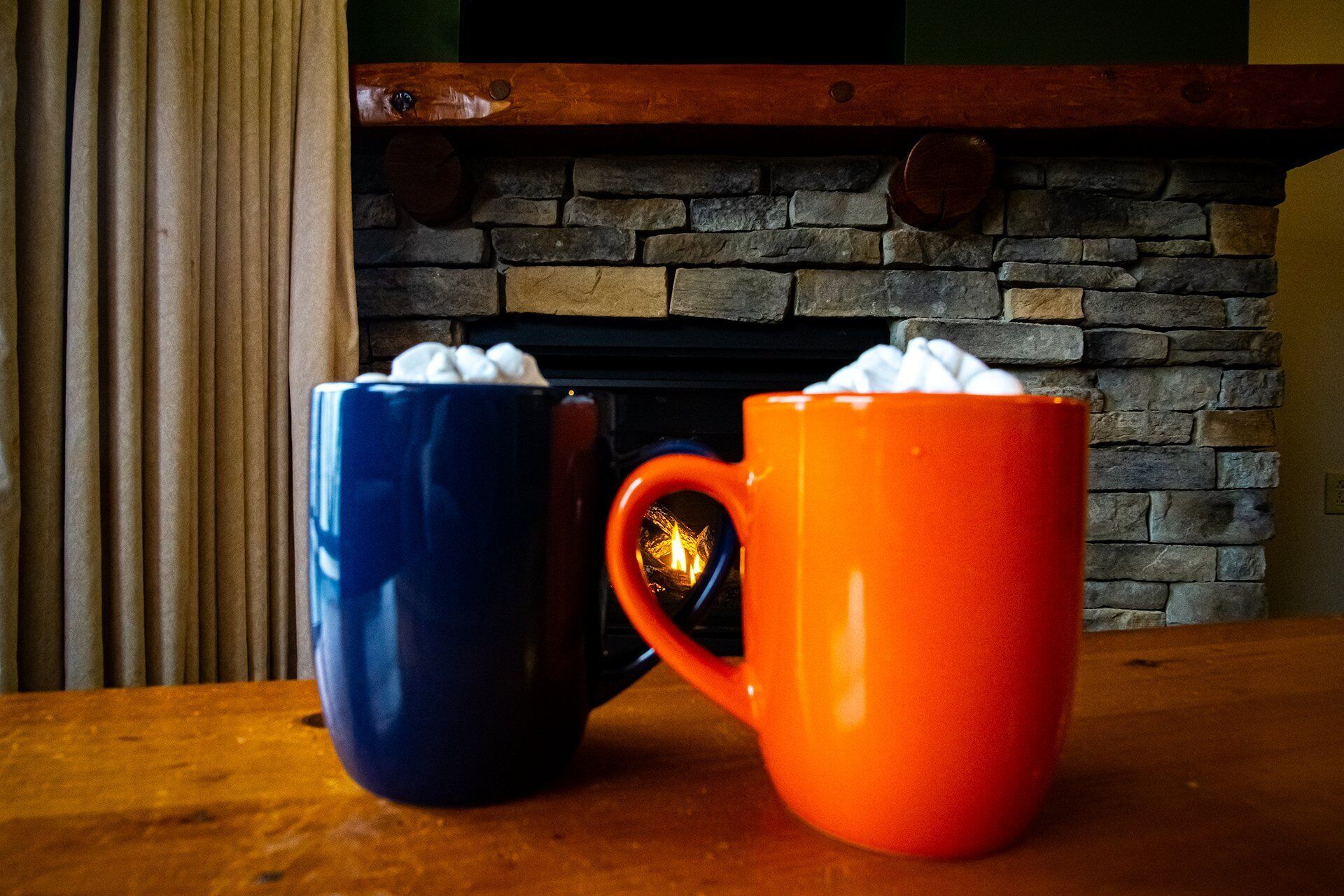 Two mugs with marshmallows on a wooden table in front of a stone fireplace.