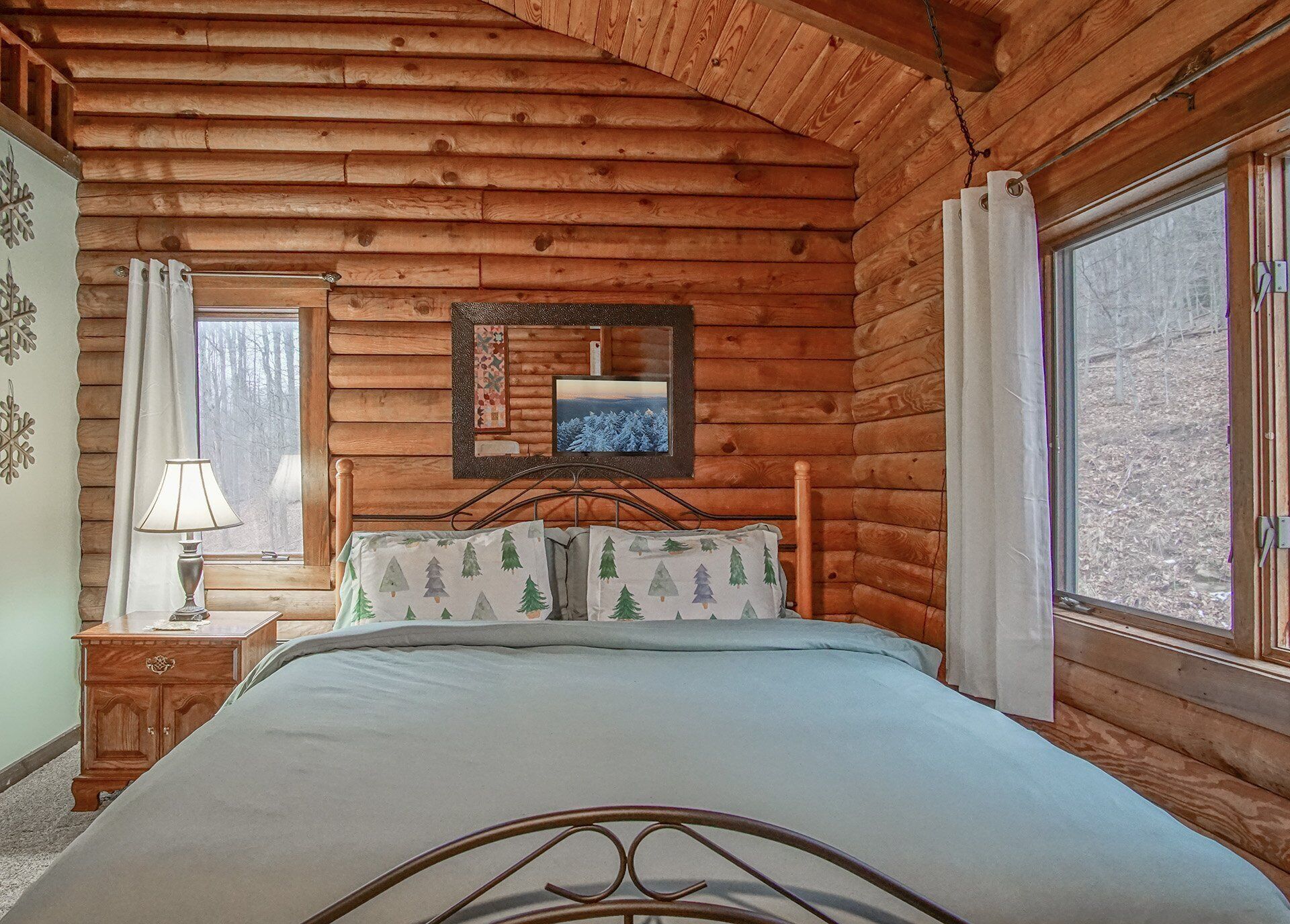A cozy log cabin bedroom with a bed, a nightstand, a lamp, a framed picture, and a window with curtains.