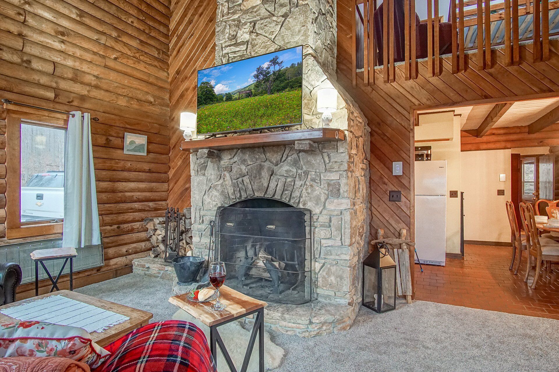 Interior acogedor de una cabaña con chimenea de piedra, paredes de madera, un televisor montado encima y una zona de comedor con mesa y sillas.