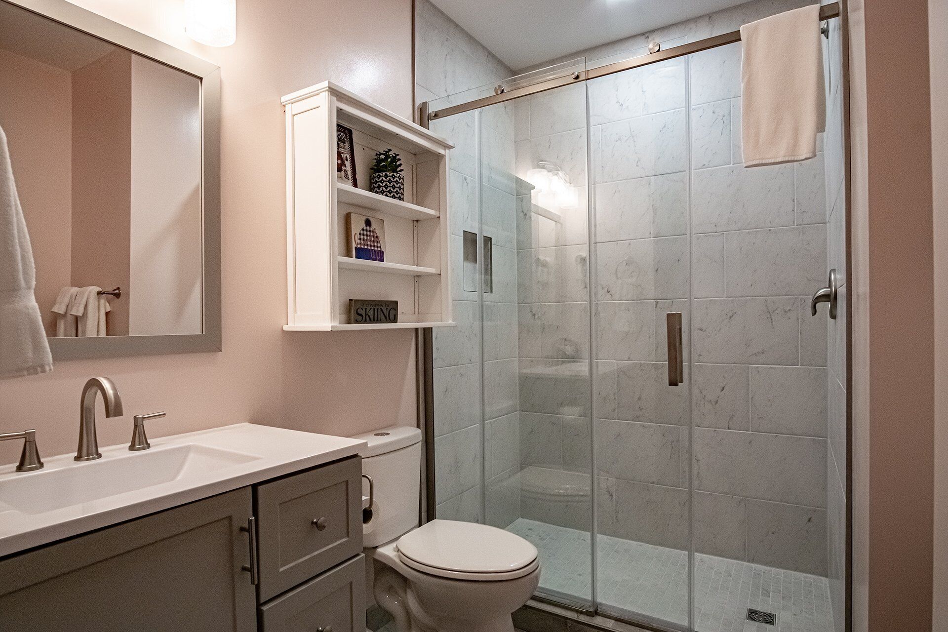 A bathroom with a white sink, a toilet, a glass-enclosed shower, and a shelf with decorative items.