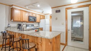 Una cocina con gabinetes de madera, encimera de granito, microondas, estufa y refrigerador. Hay tres taburetes de bar y una pequeña isla con un cuenco de fruta.