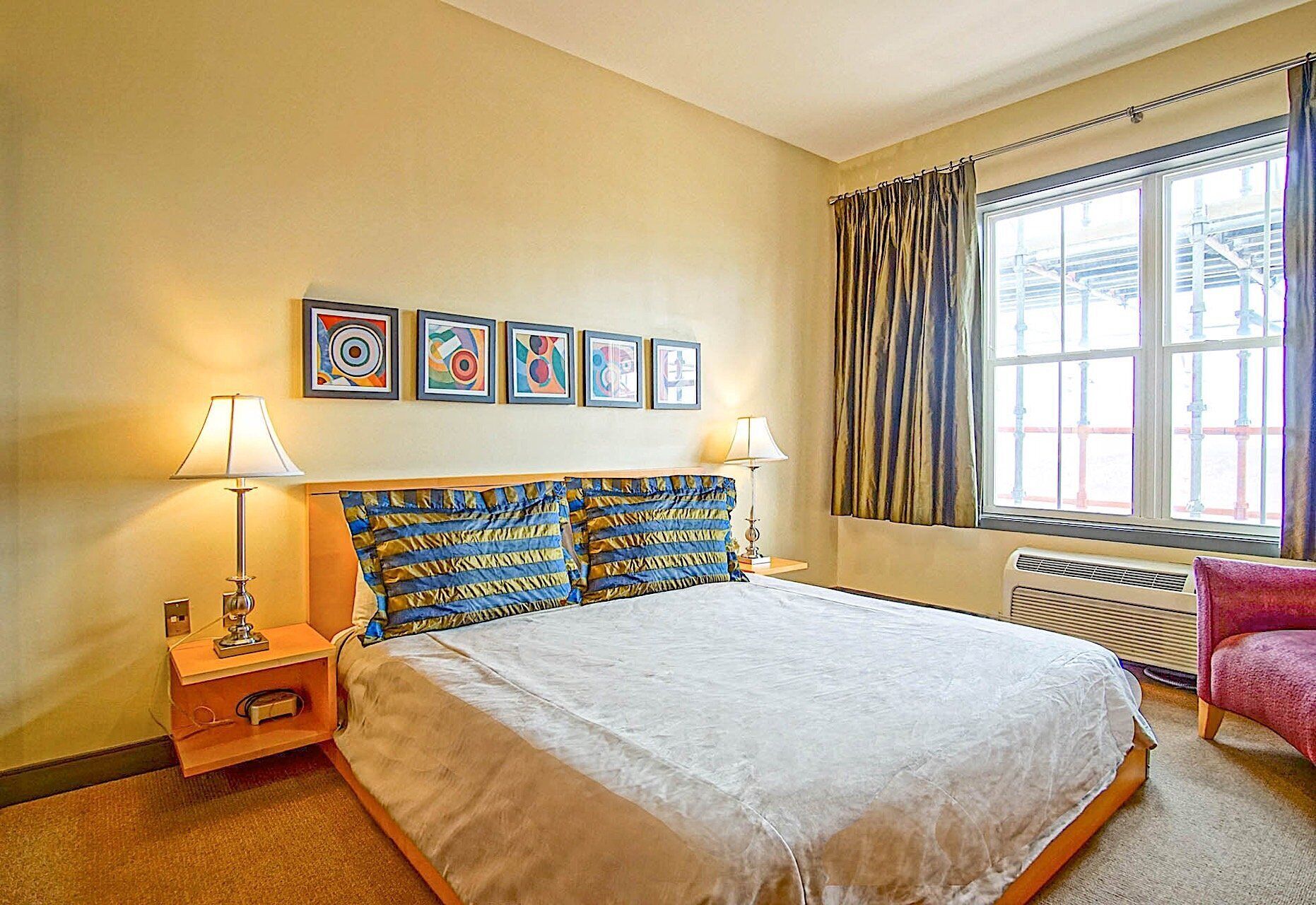 A hotel room with a bed, two bedside tables, a chair, a window with curtains, and five framed artworks on the wall.