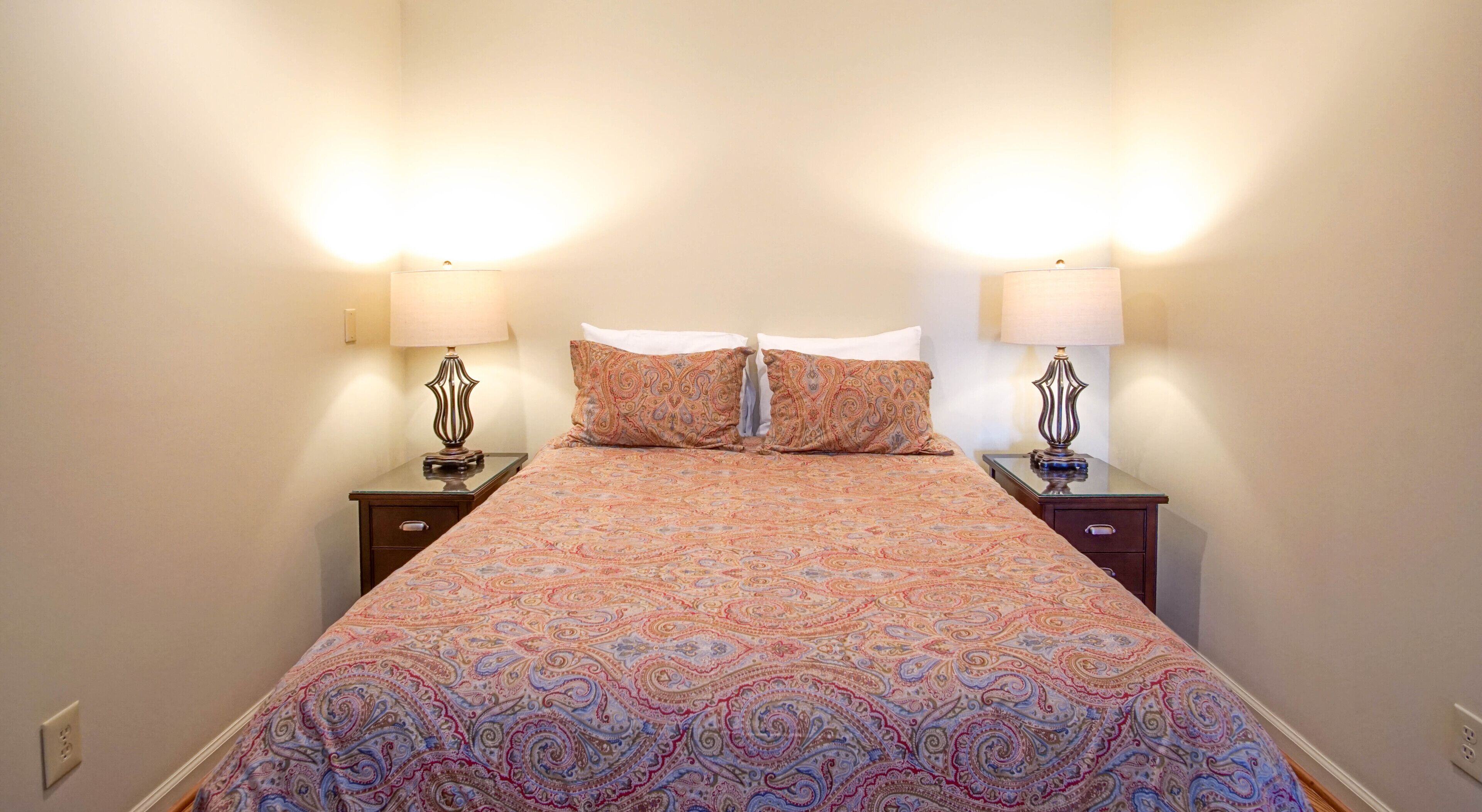 Una cama bien tendida con una colcha estampada, flanqueada por dos mesitas de noche con lámparas, en una habitación sencilla con paredes lisas.