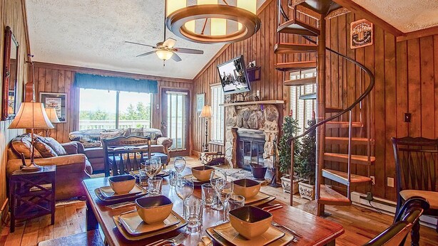 A cozy, wood-paneled room with a dining table set for four, a fireplace, a spiral staircase, and a large window offering a view of the outdoors. - Wabasso 10 Ski Hideaway Outside Your Door 2 Br Spacious Balcony (Snowshoe)