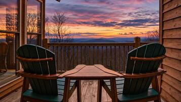 A wooden deck with two green Adirondack chairs and a wooden table, overlooking a scenic sunset view.