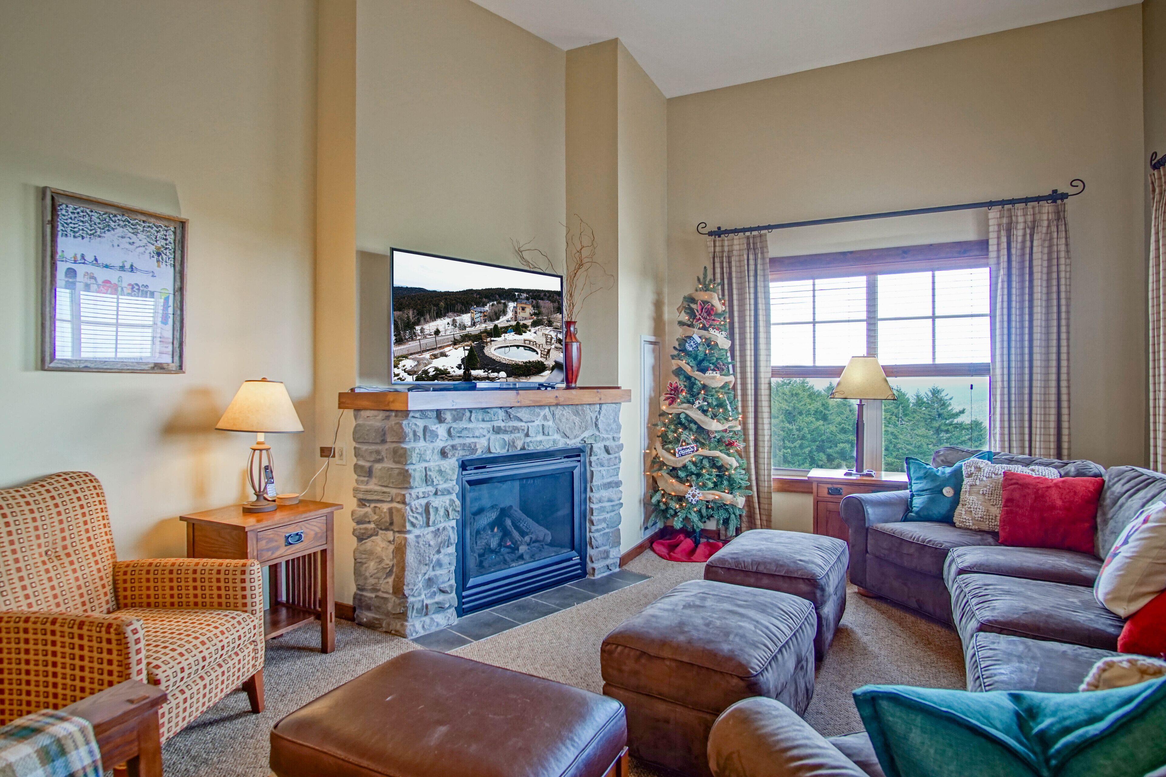A cozy living room with a stone fireplace, a television mounted above it, and a Christmas tree. There are two sofas and a coffee table.