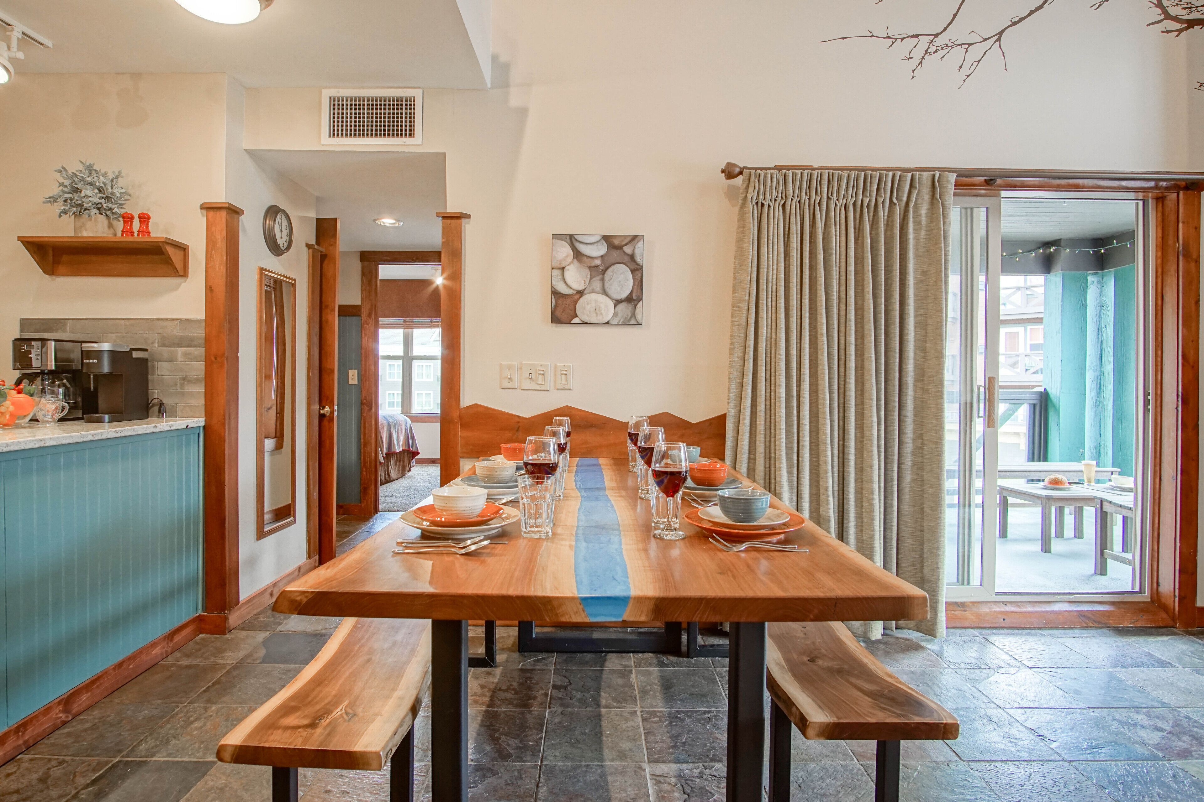 A dining area with a wooden table set for a meal, benches, a kitchen area with appliances, and a view through a glass door to an outdoor seating area.
