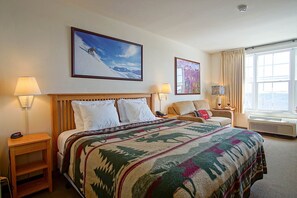Habitación de hotel con una cama, dos mesitas de noche, un sofá, una ventana con cortinas y dos cuadros colgados en la pared.
