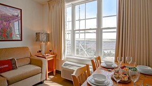 Un salon chaleureux avec un canapé, une table à manger dressée pour un repas, et une grande fenêtre offrant une vue sur un pont et des montagnes.
