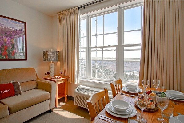 Una sala de estar acogedora con un sofá, una mesa de comedor dispuesta para una comida y un ventanal que ofrece vistas a un puente y montañas.