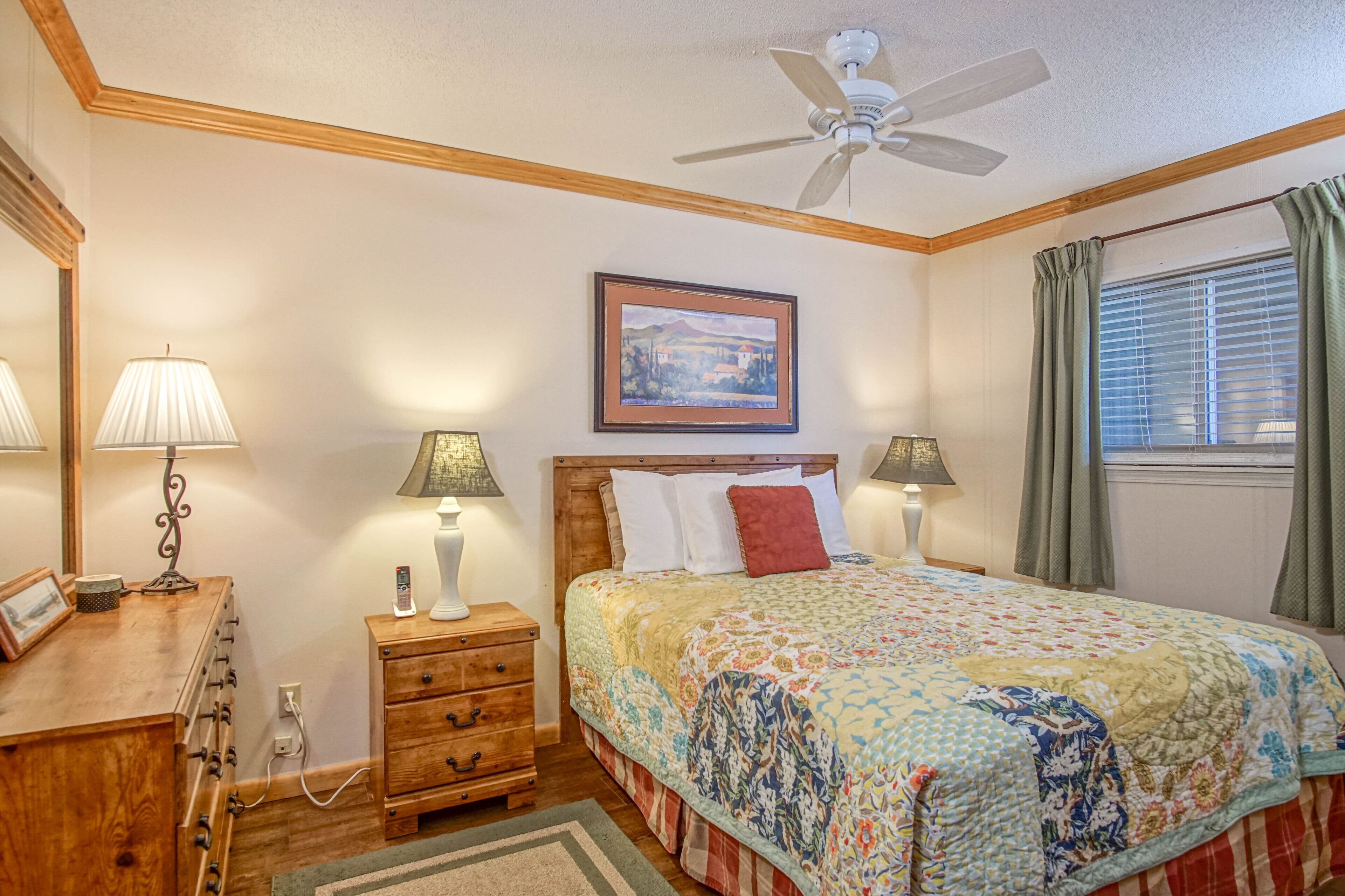 Quarto com cabeceira de madeira, cama com colcha colorida, duas luminárias de cabeceira, ventilador de teto, quadro na parede e janela com cortinas.