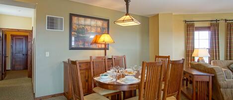 Un coin repas avec une table ronde dressée pour six personnes, entourée de chaises en bois dotées d’assises rembourrées. Un luminaire suspendu se trouve au-dessus de la table, et un tableau est accroché au mur, à gauche.