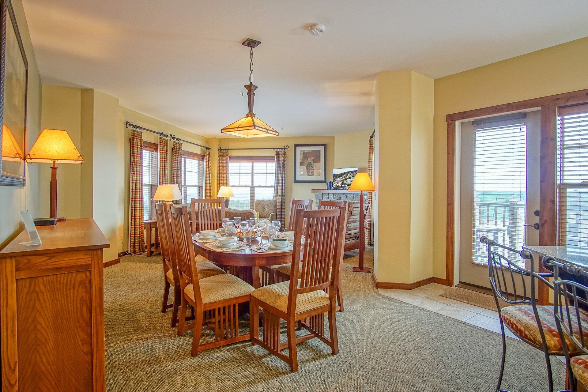 Comedor con una mesa redonda dispuesta para seis personas, rodeada de sillas de madera con asientos acolchados. Hay dos lámparas sobre la mesa y un candelabro en el techo. La habitación tiene ventanas grandes con cortinas, y una puerta que da a un balcón exterior.