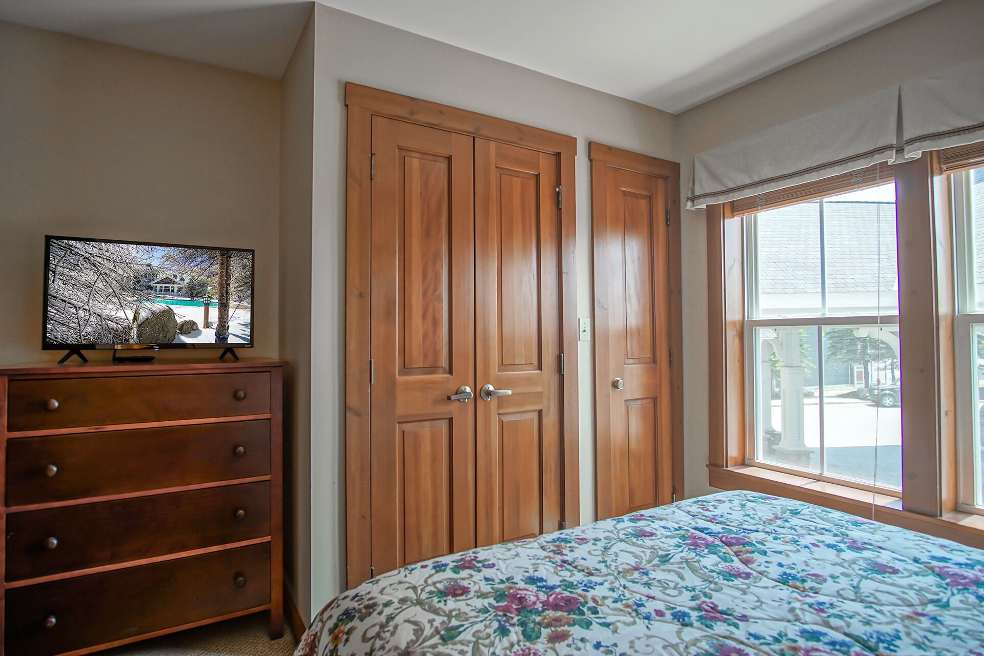 Une chambre à coucher avec une armoire en bois, une commode, un lit recouvert d’une couvre-lit à motifs floraux et un téléviseur posé sur un meuble.